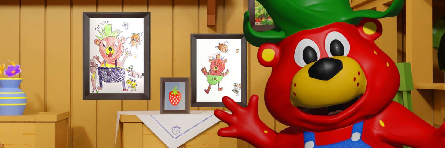 Eine Erdbeerfigur steht vor einer Holzwand mit Zeichnungen von Kindern.
